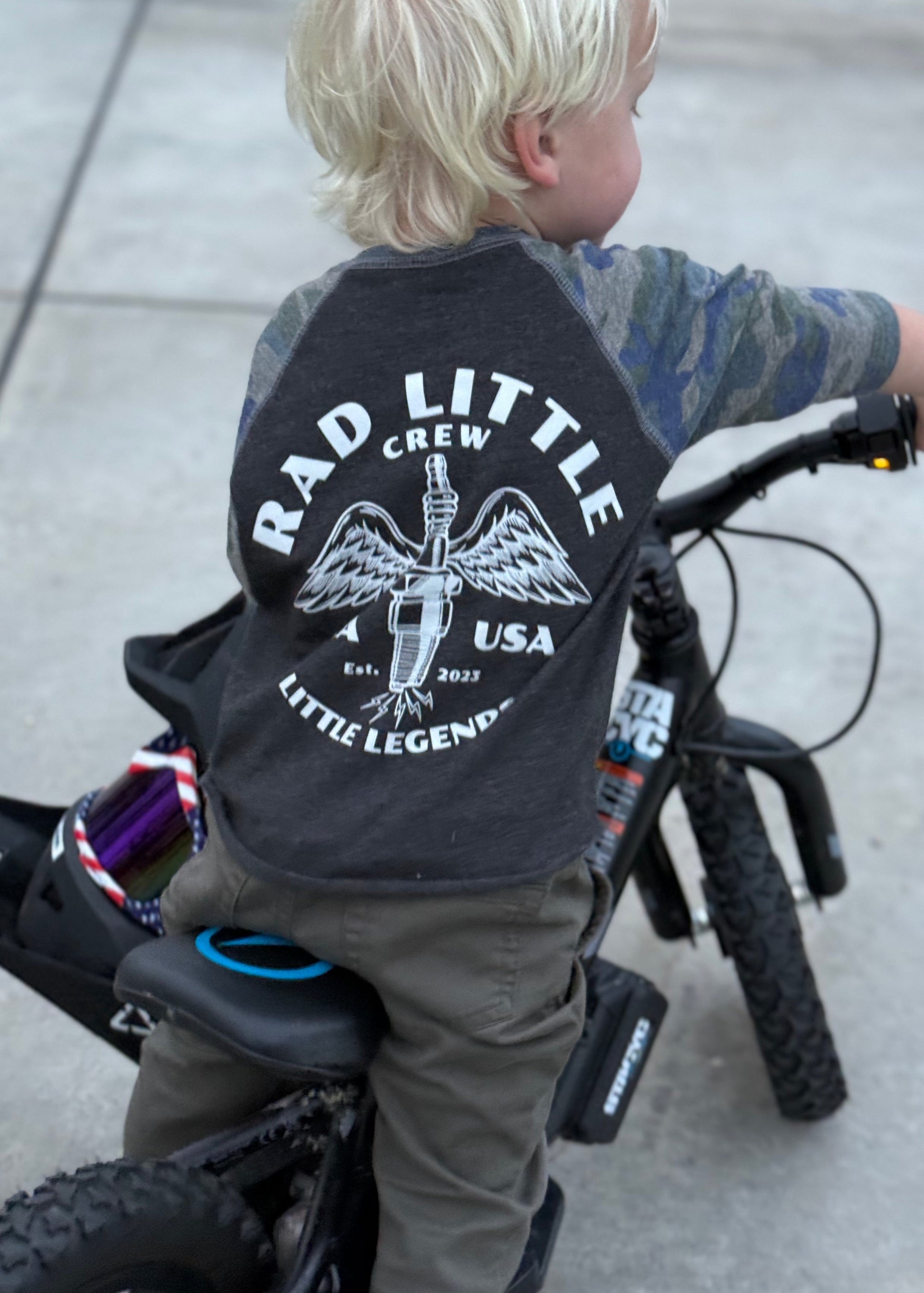 Child wearing a black t-shirt with 'Rad Little Crew' text on a bicycle.