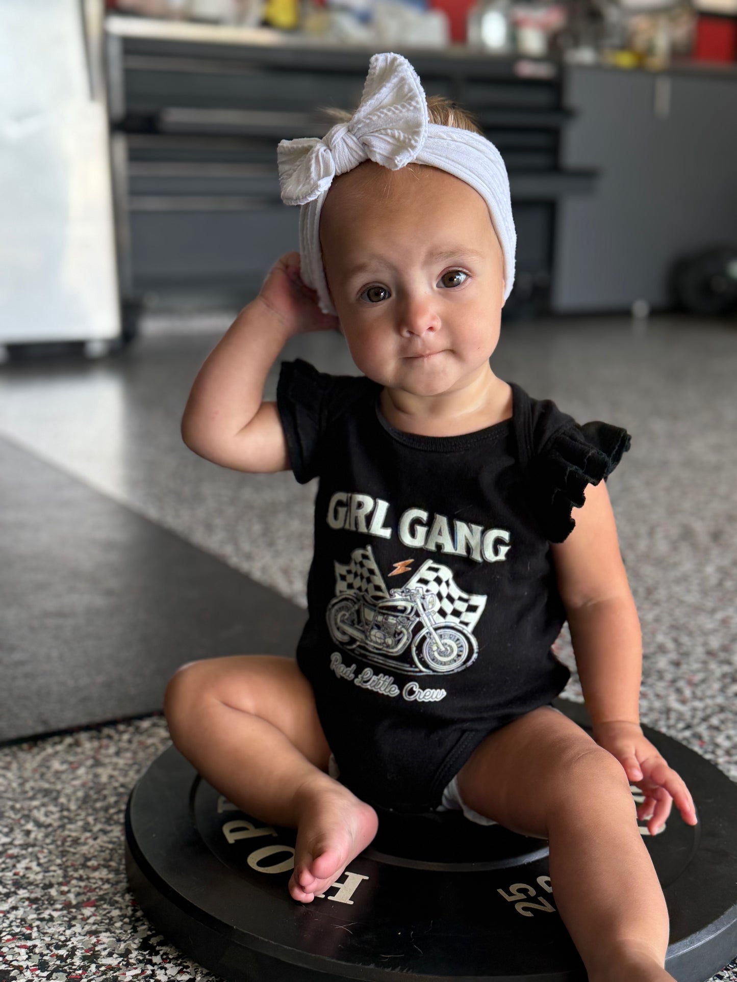 Baby wearing a black outfit with 'GRL GANG' text and graphics, sitting on a round black object.