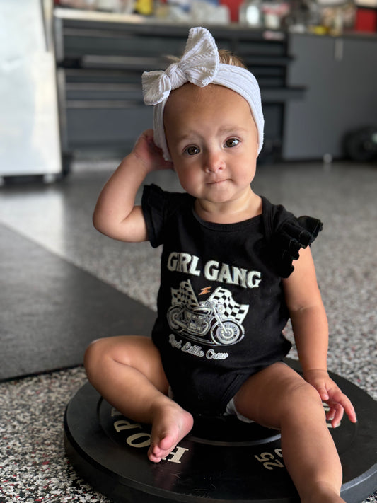 Baby wearing a black outfit with 'GRL GANG' text and graphics, sitting on a round black object.